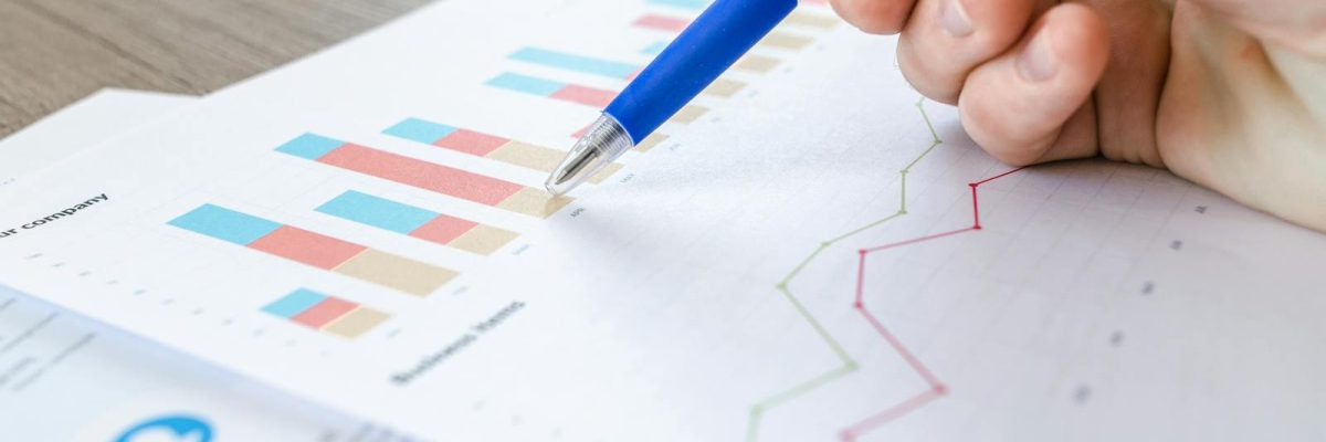A close-up of a hand with a pen analyzing data on colorful bar and line charts on paper.